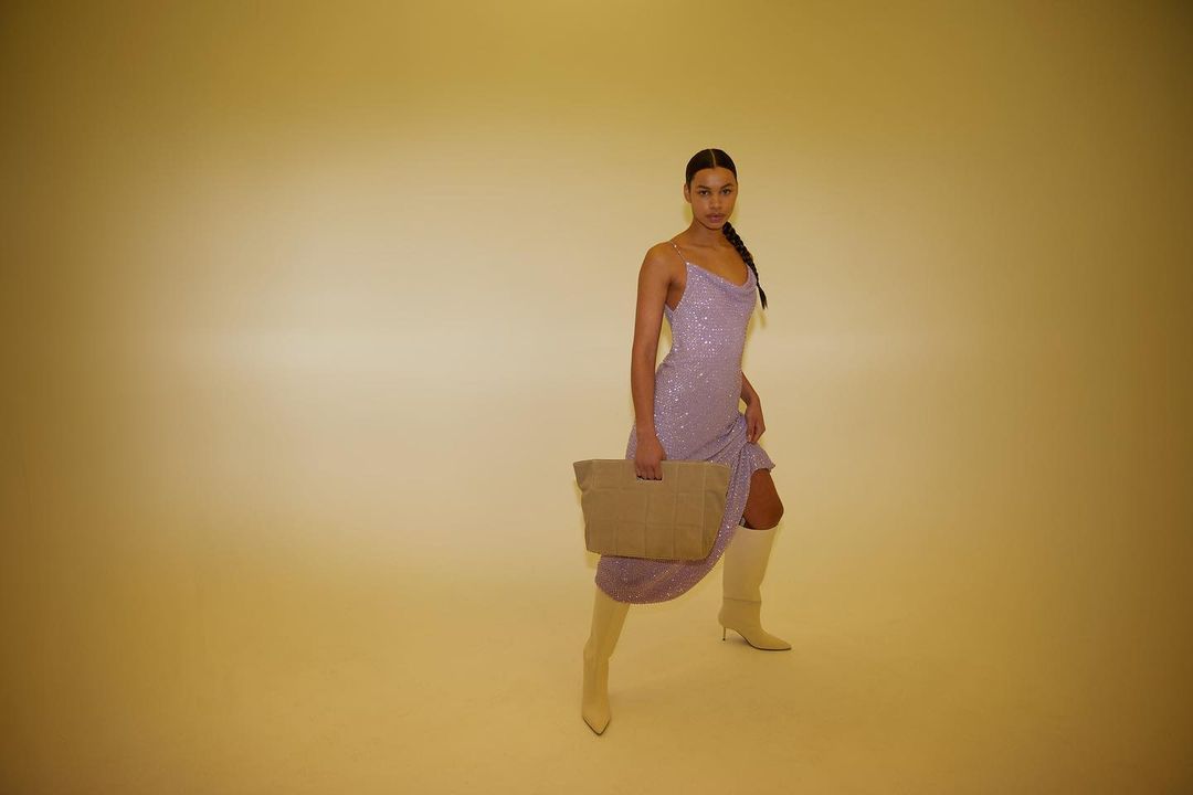 A woman in a lilac dress and beige boots poses in a studio with a tan bag, looking at the camera with a neutral expression.