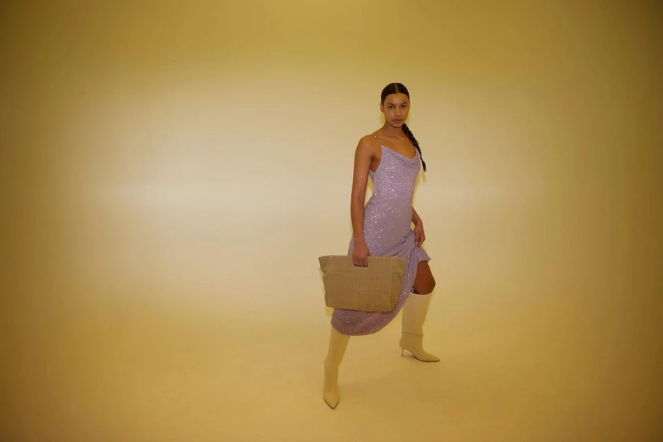 A woman in a lilac dress and beige boots poses in a studio with a tan bag, looking at the camera with a neutral expression.