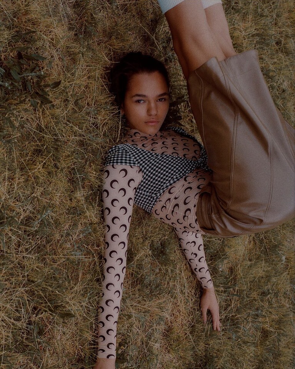 Woman in patterned top and tan pants lies on her back in grassy field in an eye-level, medium shot, in outdoor fashion editorial.