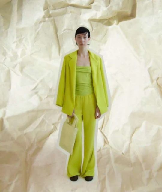 A slender woman poses in a vibrant chartreuse suit and matching top against a textured, off-white backdrop, carrying a light yellow handbag.