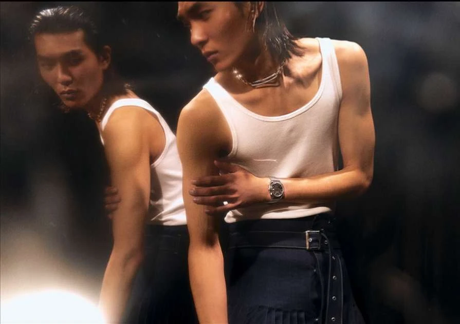 Fashion shot of two adult male models wearing white tank tops and dark pleated skirts against a dark backdrop, studio setting.