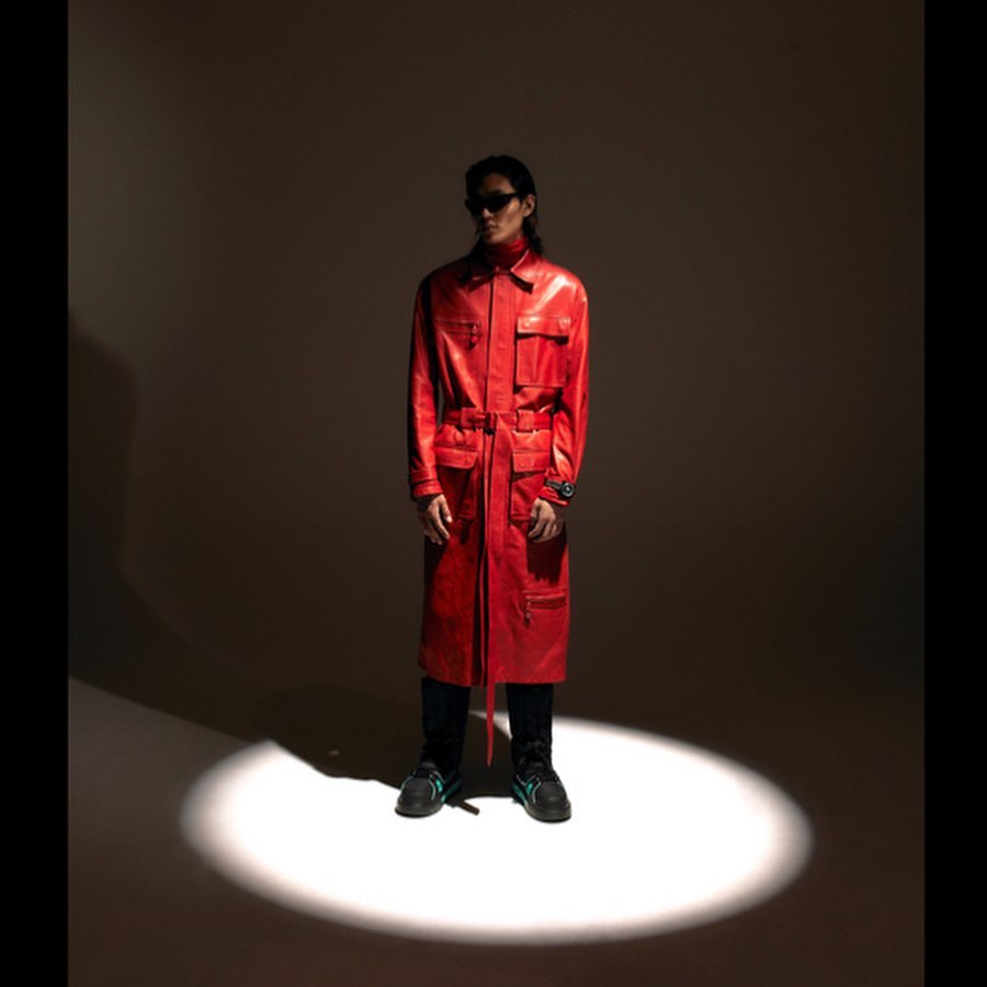 A stylish adult male poses in a long red leather coat and black sunglasses in a spotlight against a dark background, showcasing a studio fashion shot.