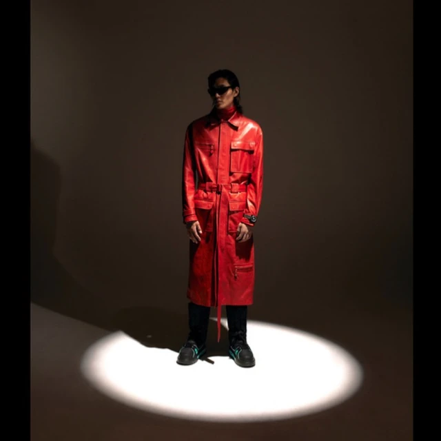 A stylish adult male poses in a long red leather coat and black sunglasses in a spotlight against a dark background, showcasing a studio fashion shot.
