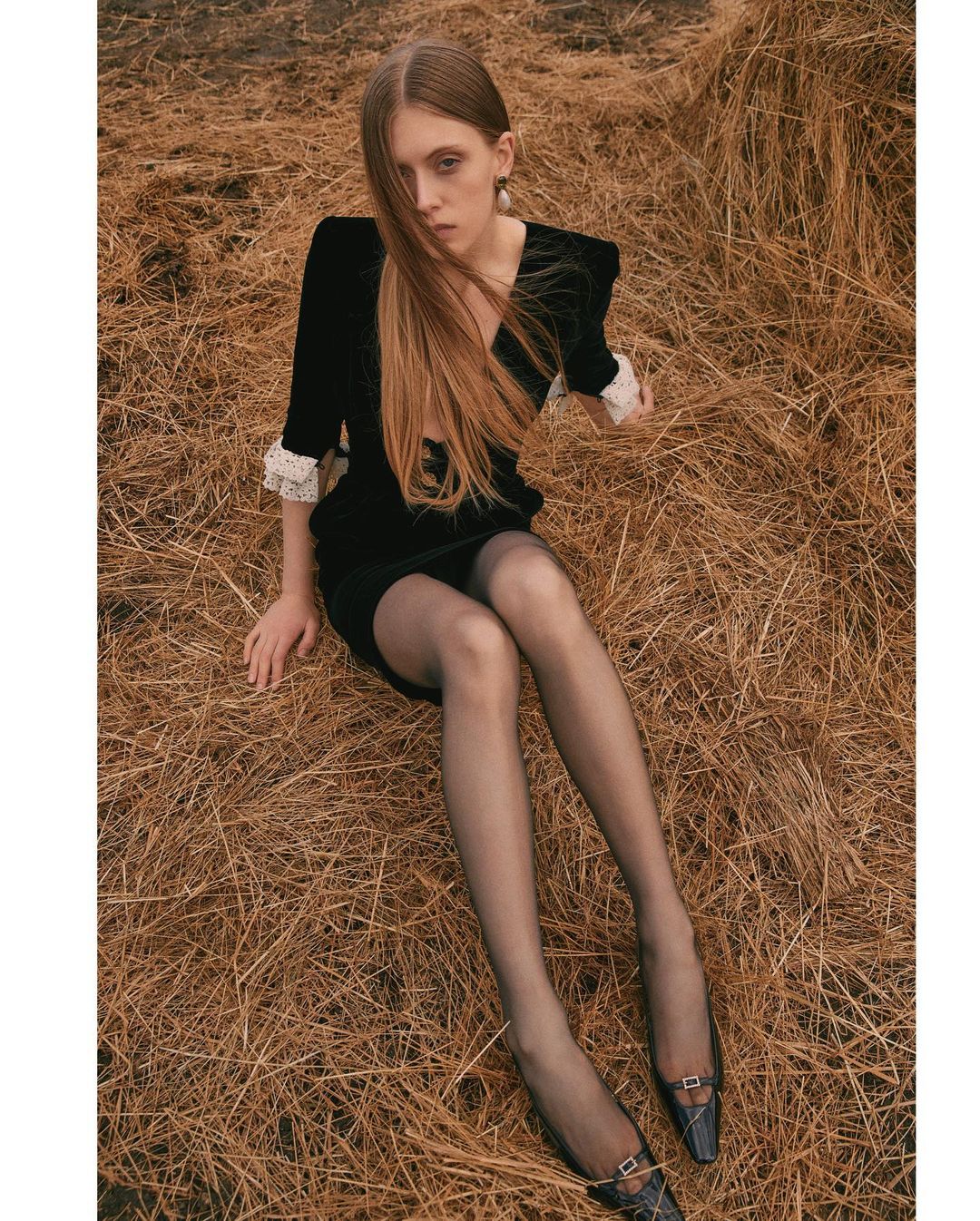 Model seated on a bed of hay wearing a black dress with white cuffs and sheer black stockings