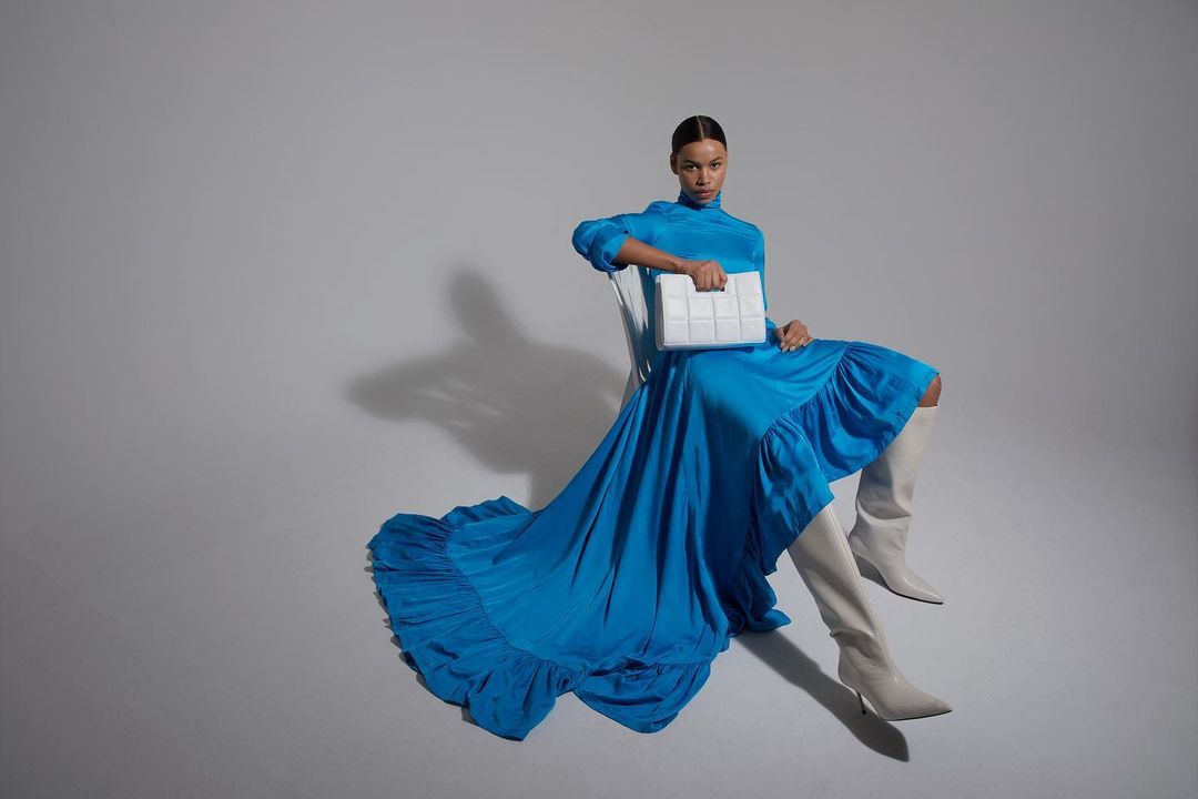 A woman in a blue gown and high boots sits holding a white bag against a light-grey background in a studio fashion shot.