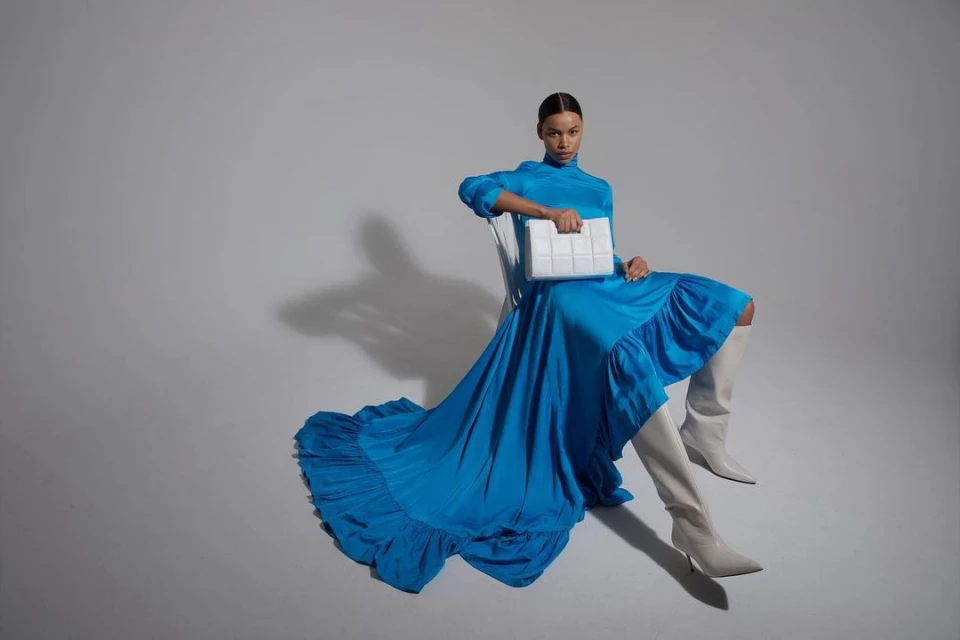 A woman in a blue gown and high boots sits holding a white bag against a light-grey background in a studio fashion shot.