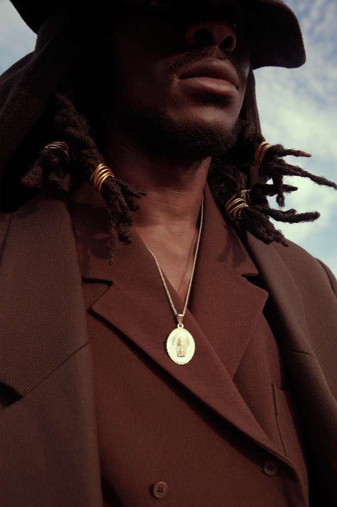 Portrait of stylish black man wearing brown coat and hat with dreadlocks and gold jewelry against a blue sky with clouds.