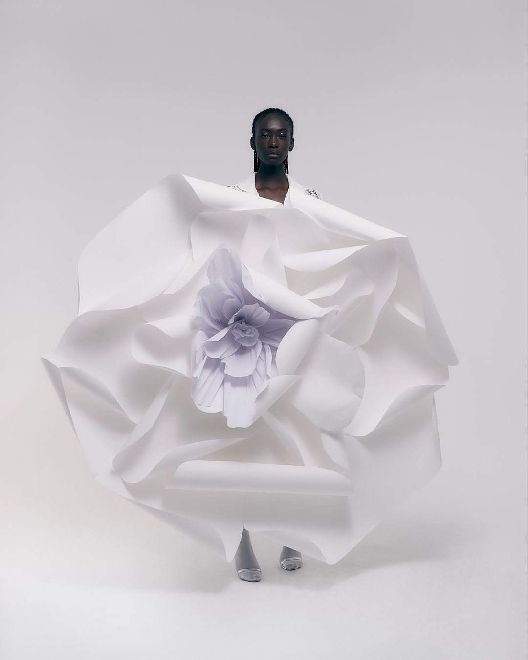 Fashion shot of a Black female model wearing a white paper dress with a large flower, posing in a white studio with soft lighting.