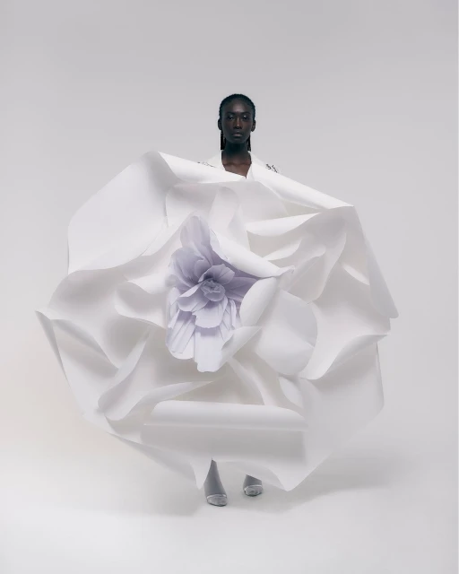 Fashion shot of a Black female model wearing a white paper dress with a large flower, posing in a white studio with soft lighting.