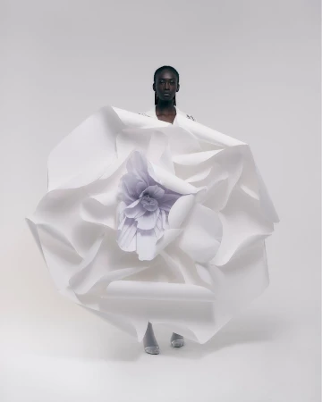 Fashion shot of a Black female model wearing a white paper dress with a large flower, posing in a white studio with soft lighting.
