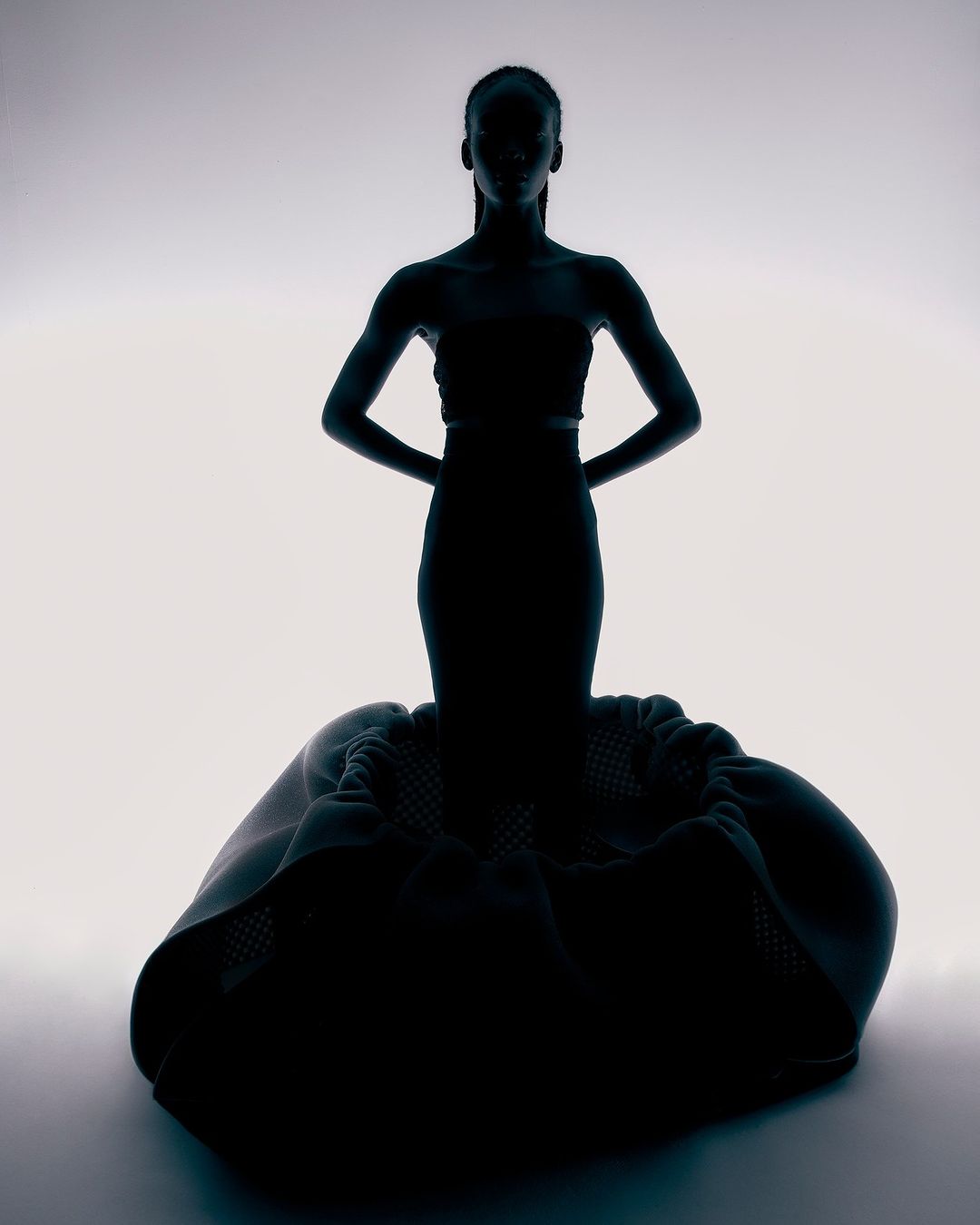 Silhouette of a slender adult woman in a modern black gown, standing in a white studio, hands behind her back, creating a fashion-forward look.