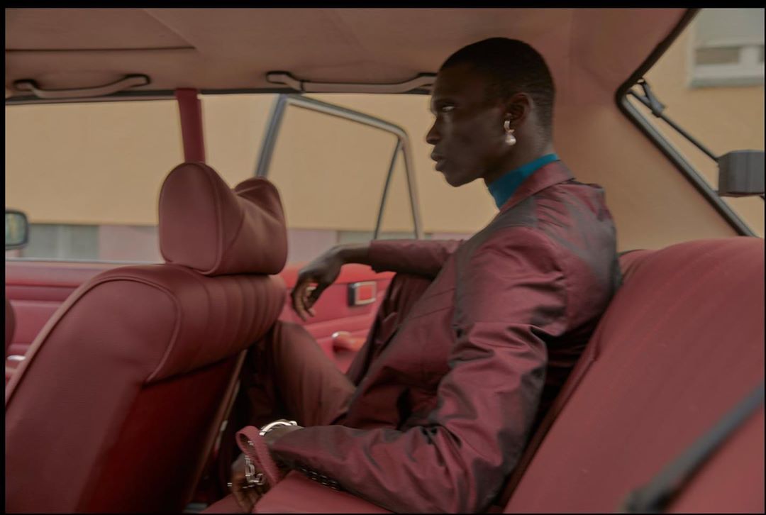 Stylish adult in burgundy suit sitting in vintage car with red interior, wearing pearl earring, looking out the open window.