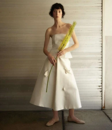 A woman in a white dress holds a yellow flower against a neutral garage door, creating a minimalist yet striking fashion editorial.
