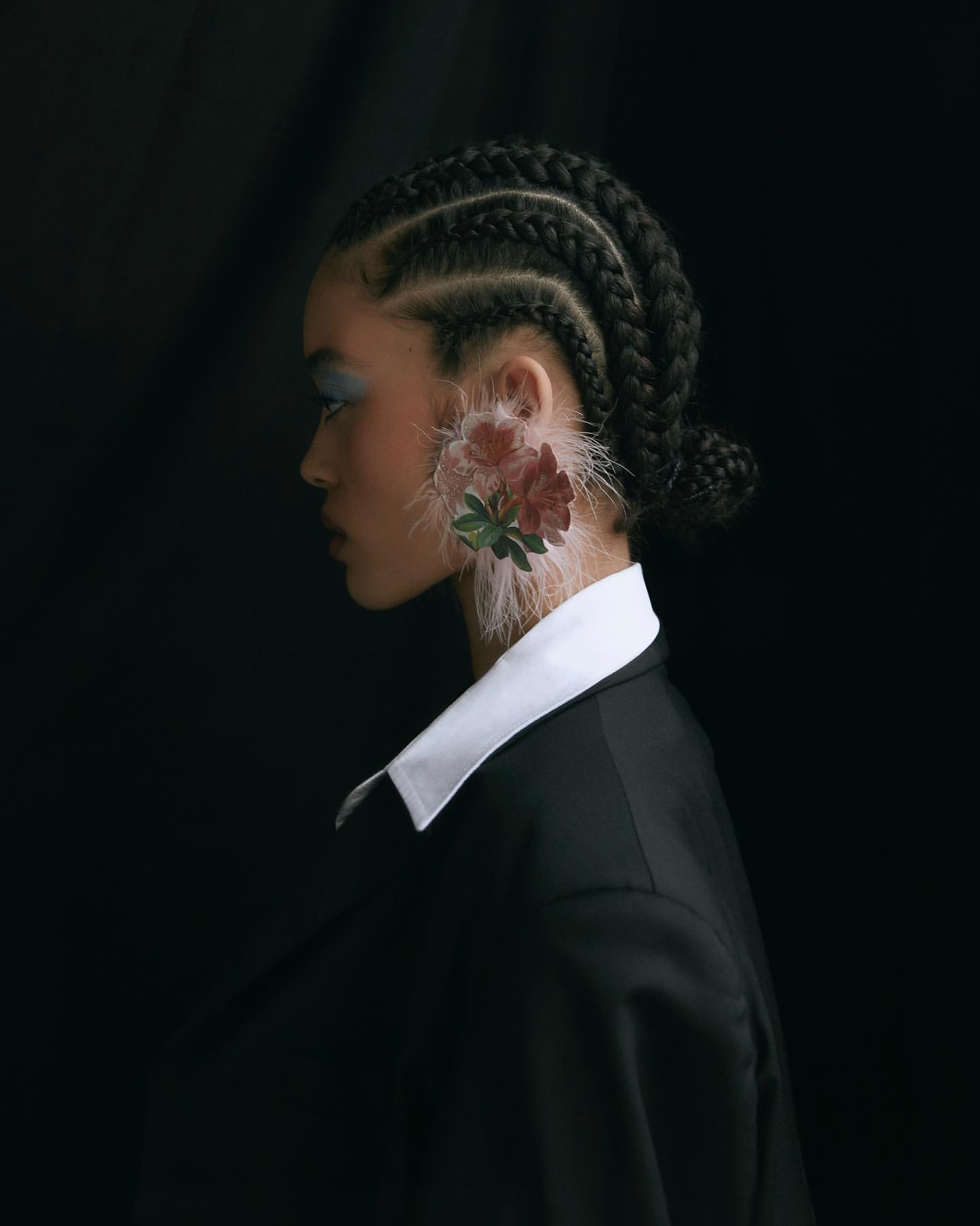 Profile portrait of a woman with braided hair and elaborate flower and feather ear decoration against a dark backdrop in a studio setting.