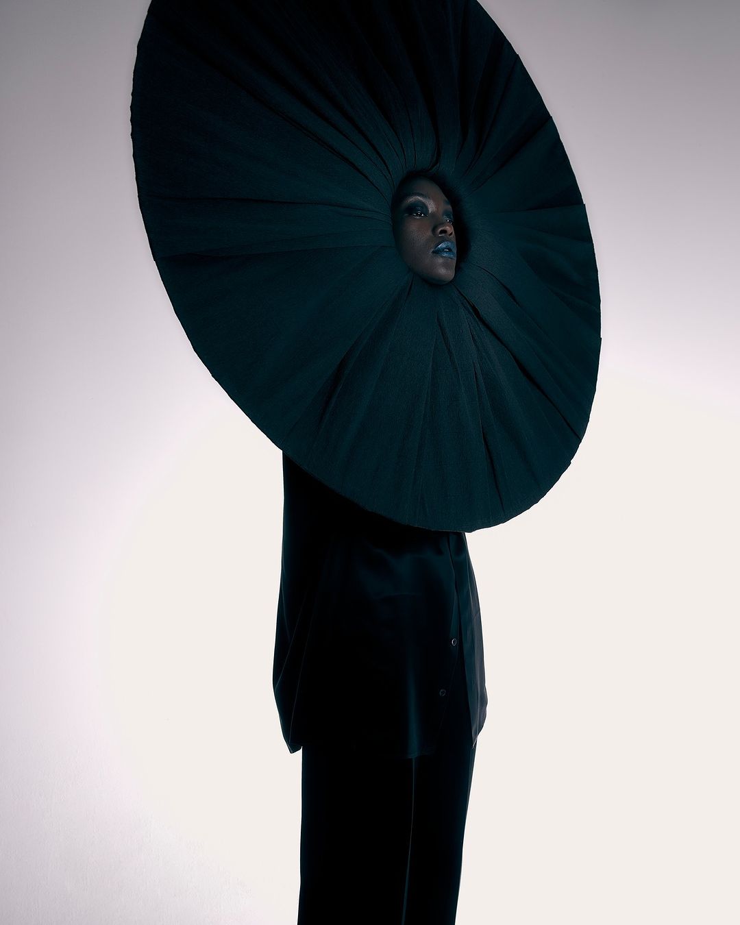 Fashion-forward woman poses in dramatic, round, dark-green couture garment against a bright studio backdrop for a striking, avant-garde portrait.