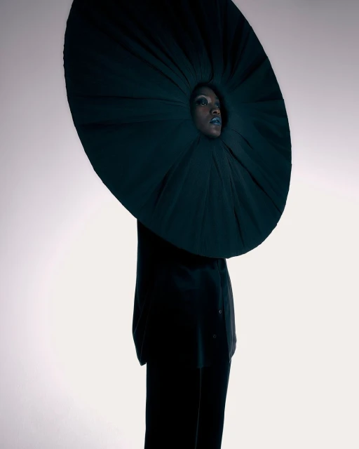 Fashion-forward woman poses in dramatic, round, dark-green couture garment against a bright studio backdrop for a striking, avant-garde portrait.