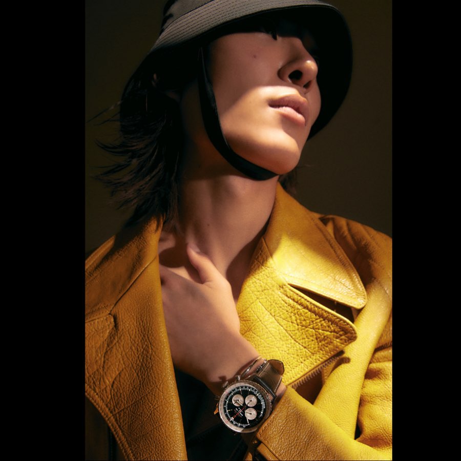 Close-up shot of an adult with a bucket hat and yellow leather jacket. Person wears a wristwatch and poses in soft lighting.