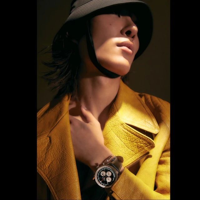 Close-up shot of an adult with a bucket hat and yellow leather jacket. Person wears a wristwatch and poses in soft lighting.