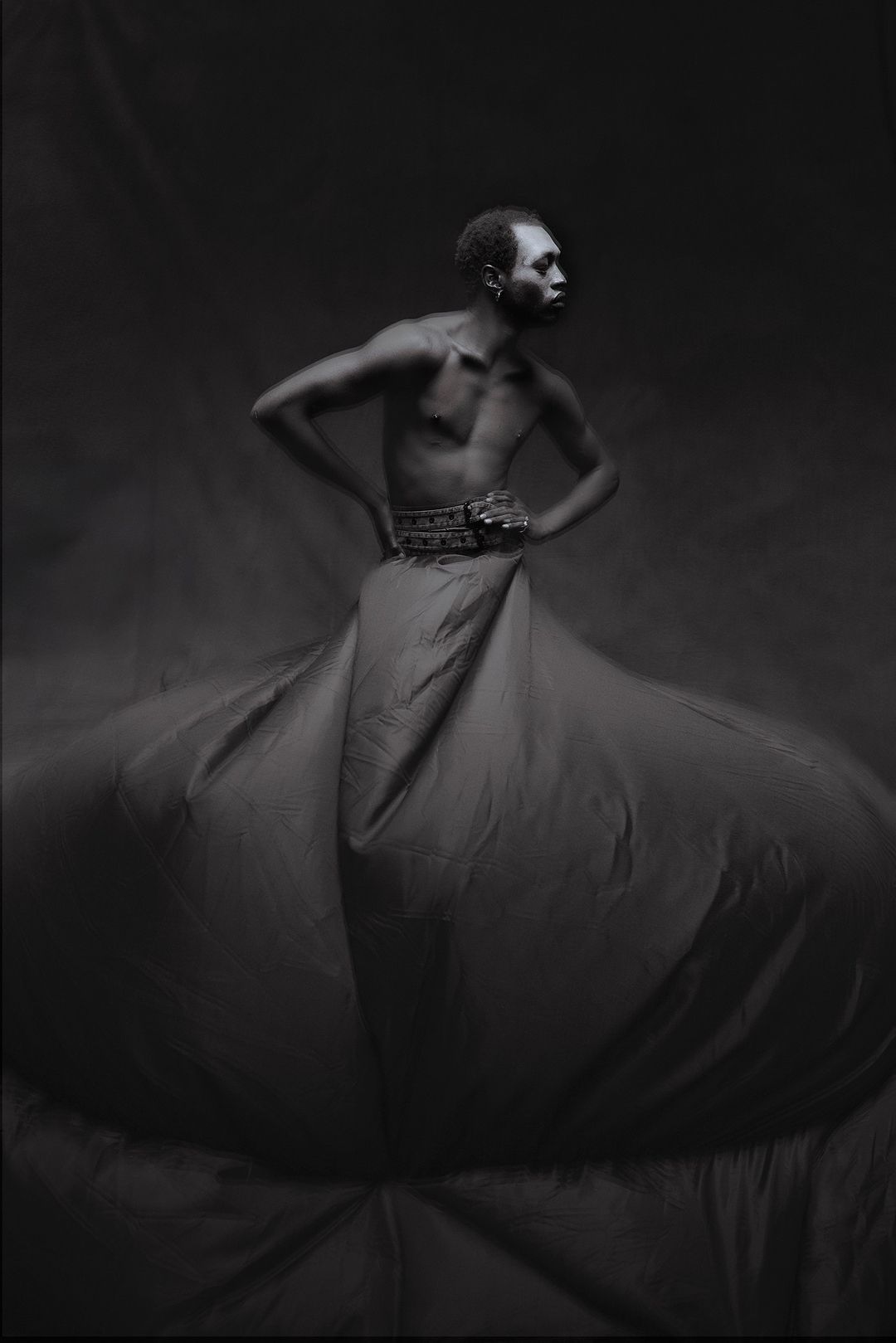 Dramatic black and white portrait of Black male model with hands on hips, wearing decorative belt and large fabric skirt in studio.