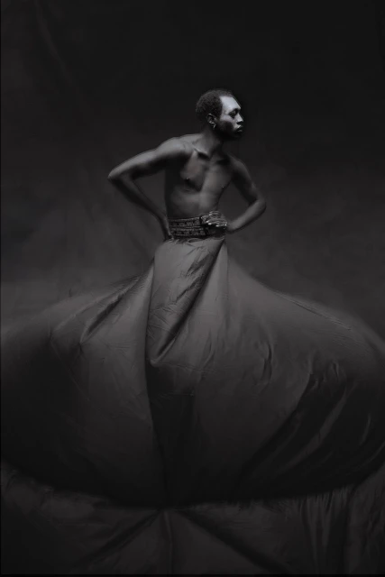 Dramatic black and white portrait of Black male model with hands on hips, wearing decorative belt and large fabric skirt in studio.