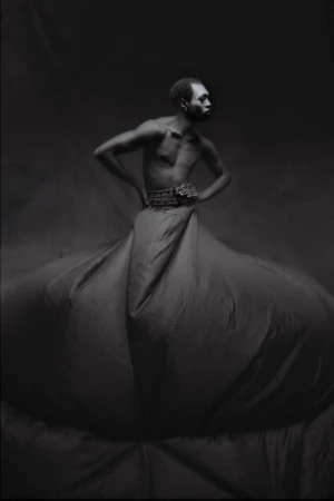 Dramatic black and white portrait of Black male model with hands on hips, wearing decorative belt and large fabric skirt in studio.