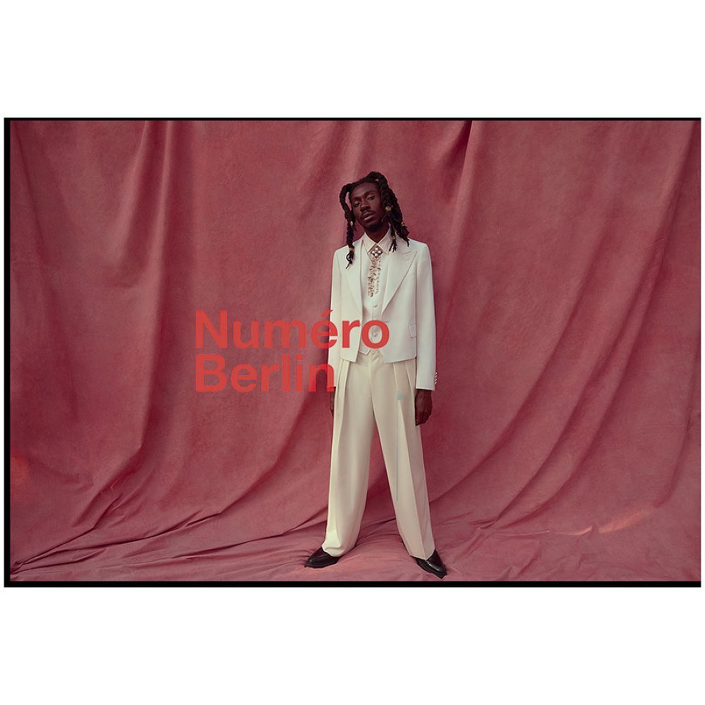 An adult male poses in an off-white suit in front of a draped red-pink textile background, with the text 'Numero Berlin' visible.