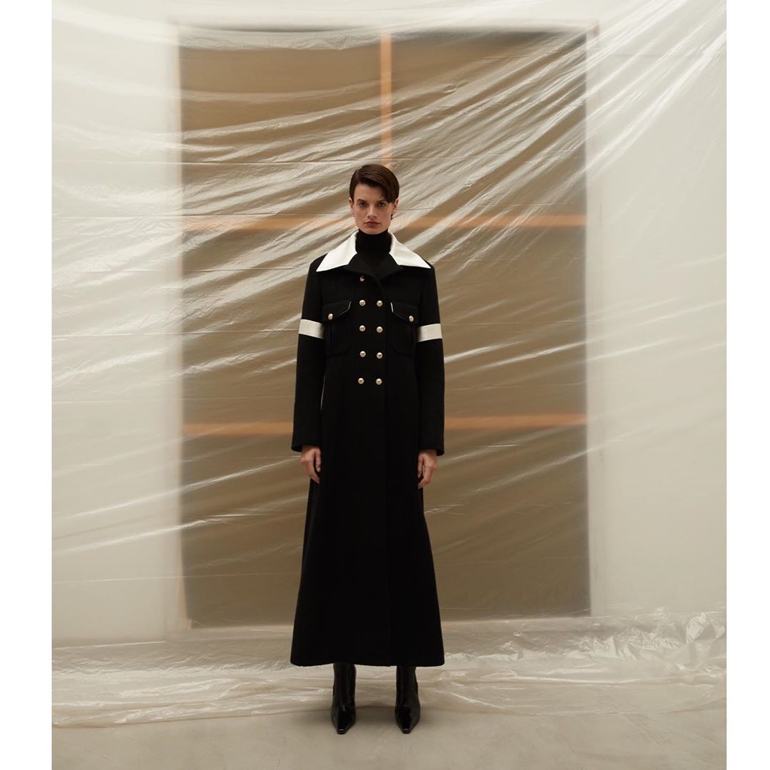 Full-length shot of a model wearing a long black coat with white accents, standing in front of a plastic-covered backdrop in a studio.