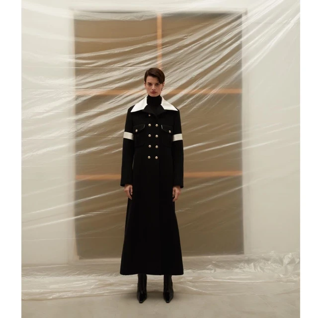 Full-length shot of a model wearing a long black coat with white accents, standing in front of a plastic-covered backdrop in a studio.