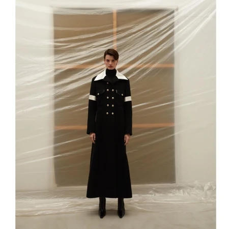 Full-length shot of a model wearing a long black coat with white accents, standing in front of a plastic-covered backdrop in a studio.