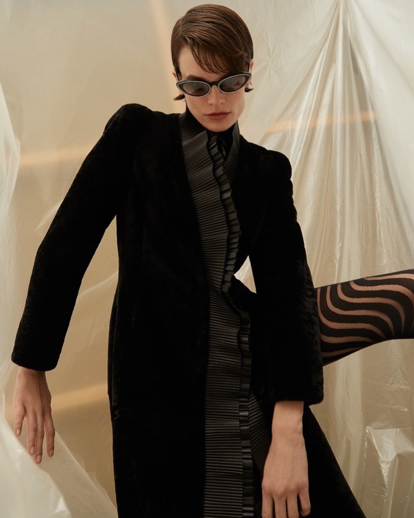 Woman in sunglasses poses wearing a black coat with gray trim against a backdrop of white plastic sheeting. Fashion editorial.