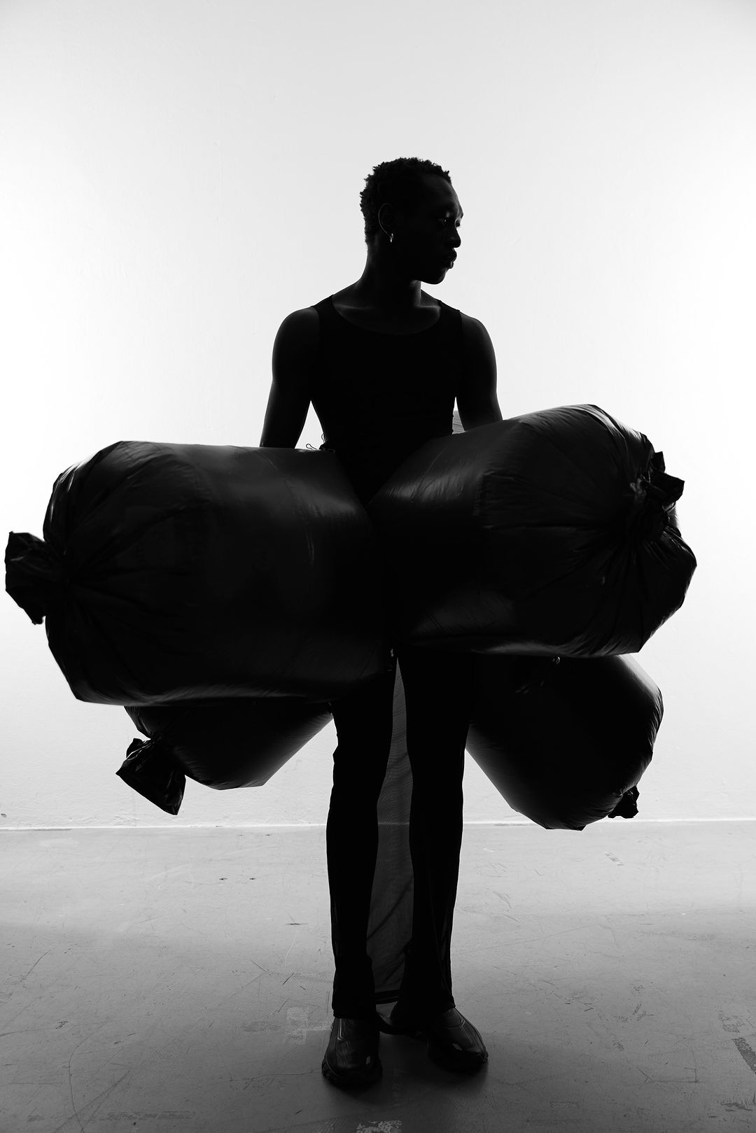 Monochrome studio shot of an adult male figure artistically posing with what appears to be filled trash bags, sculptural fashion.