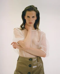 Model wearing a sheer white top and brown glossy skirt with her hand at her neck against a plain white background