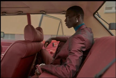 Black man in a shiny burgundy suit and blue turtleneck seated in the back of a vintage car with red leather interior.
