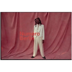 Man wearing a cream suit and white tie standing against a pink draped fabric background with red typography