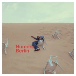 Person in blue suit lying on sand dunes surrounded by scattered white plastic chairs