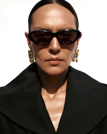 Close-up portrait of a person wearing black sunglasses and textured gold earrings against a white background