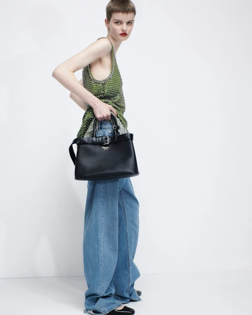 Fashion shot of a woman with short hair wearing a green beaded top, jeans, black flats, and holding a black leather handbag.