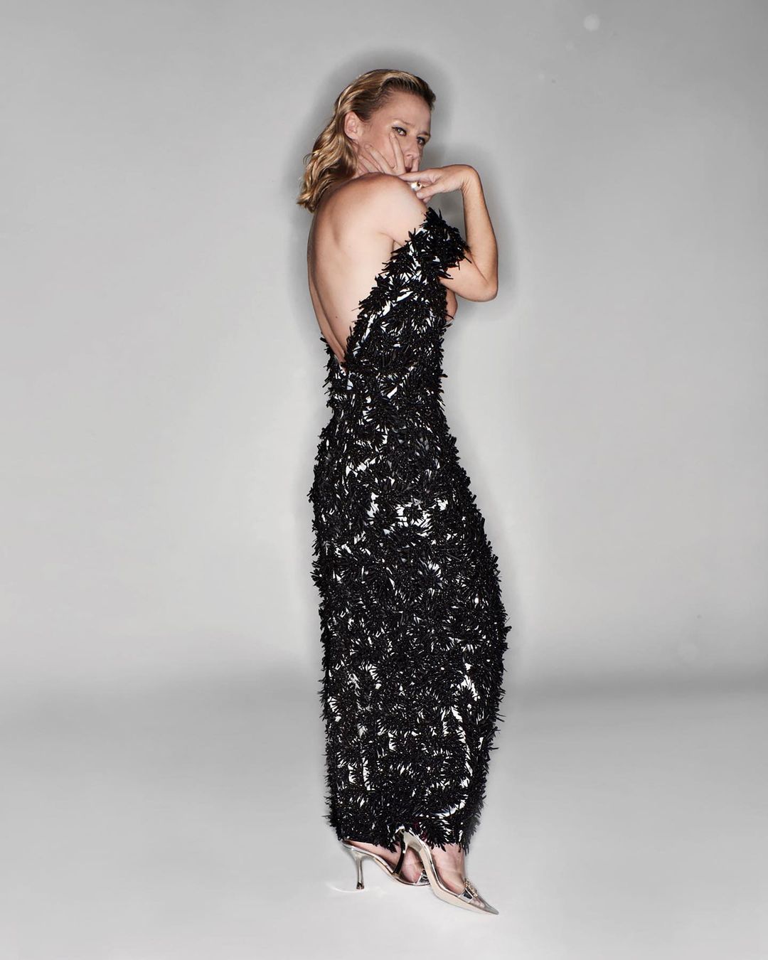 Blonde woman in a backless black gown poses in a studio with silver heels, looking over her shoulder at the camera, against a grey backdrop.
