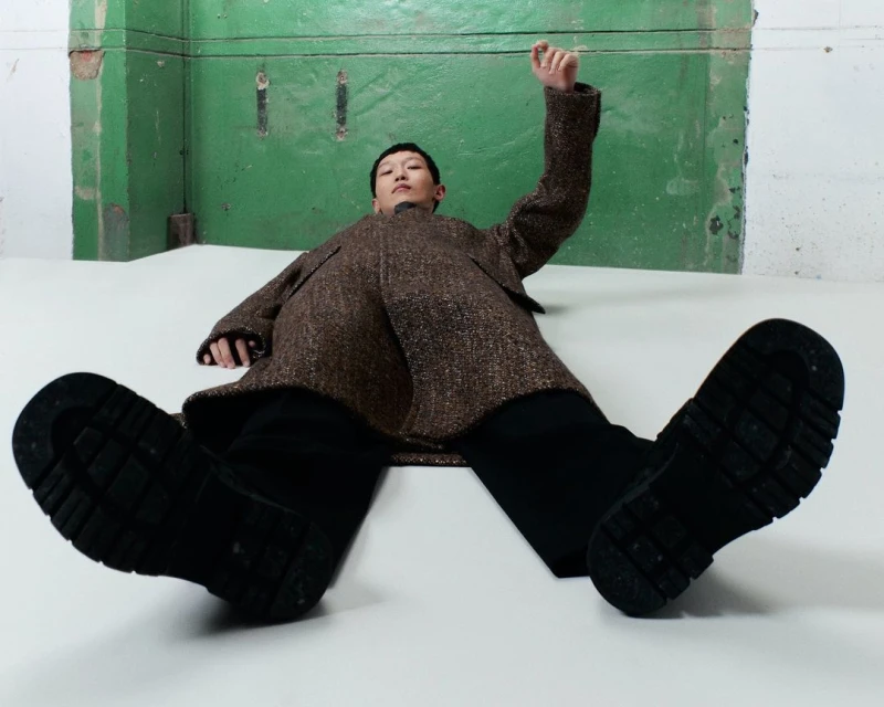 Fashion shot of a person lying on the floor wearing black boots and a coat, raising their right arm in front of a green wall.