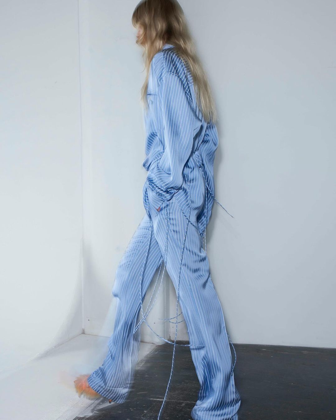 Fashion photo of a slender adult female model walking indoors, wearing a light blue striped pajama set and light orange shoes in a studio.