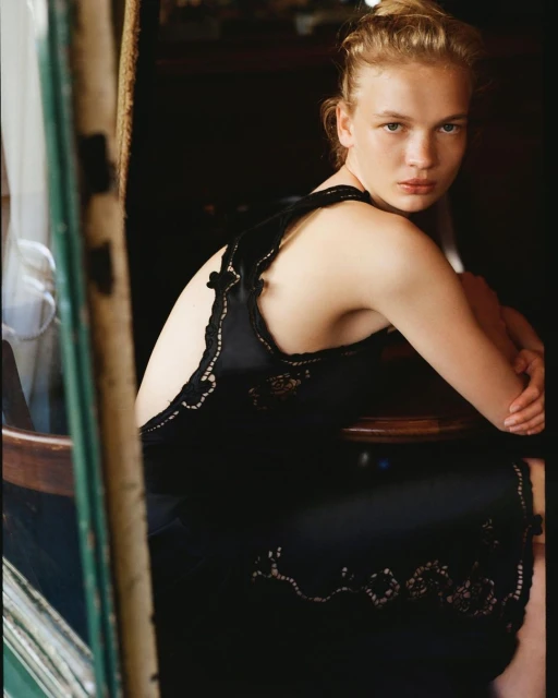 Young woman in elegant black dress with lace detail sitting indoors with soft lighting, looking at the camera, fashion photography.