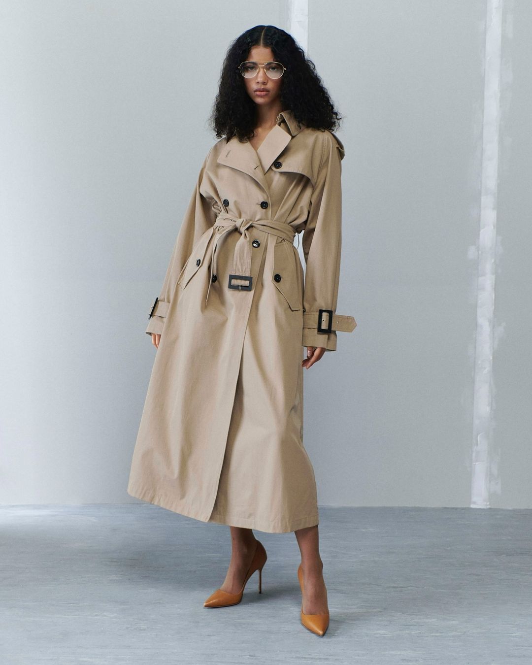 Woman in a trench coat standing in front of a grey wall. She is wearing glasses and high heels, posing in a studio setting.