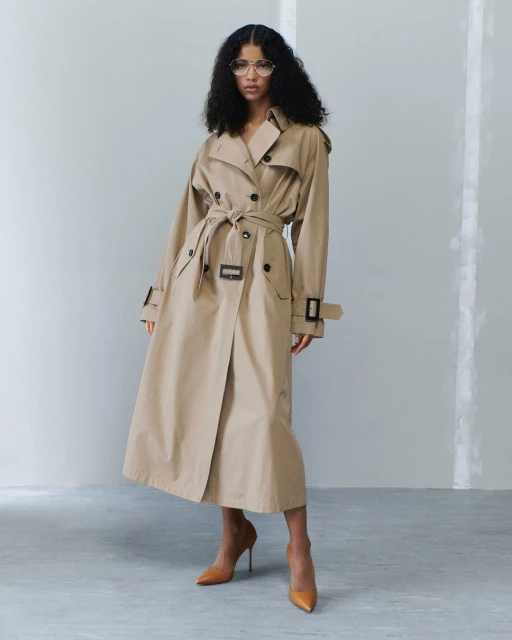 Woman in a trench coat standing in front of a grey wall. She is wearing glasses and high heels, posing in a studio setting.