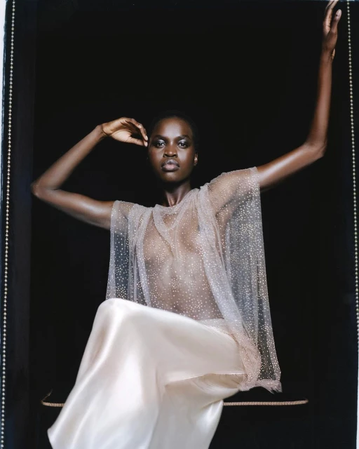 Elegant portrait of a Black woman in a sheer beaded top and satin skirt against a dark backdrop in a fashion forward pose.