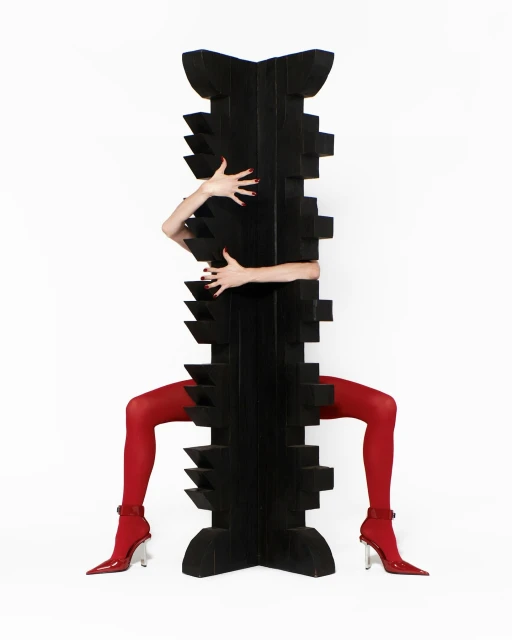 Abstract studio shot featuring red-stockinged legs in heels and outstretched arms around a geometrical black sculpture on white backdrop.
