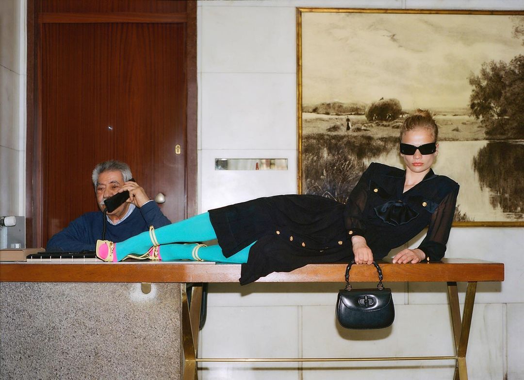 Fashion shot of a woman in black dress and sunglasses reclining on a counter, with a man on the phone and a landscape art in background.