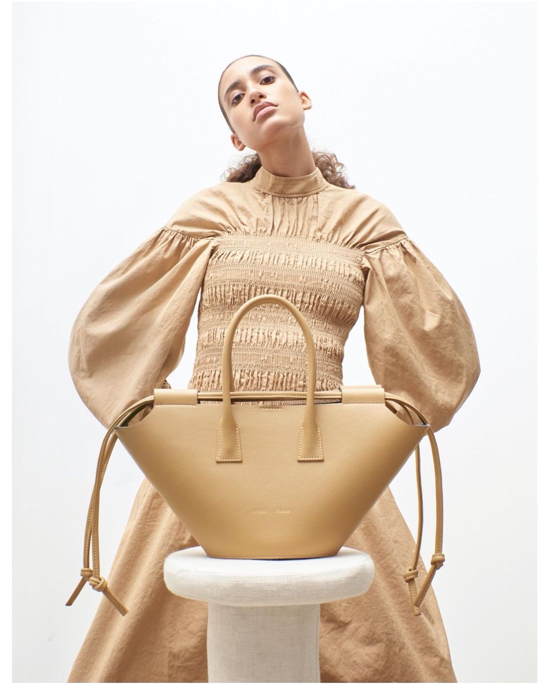 Tan-colored leather handbag sits on a white stool, woman with tan top and dress poses behind it in a studio setting, neutral lighting.