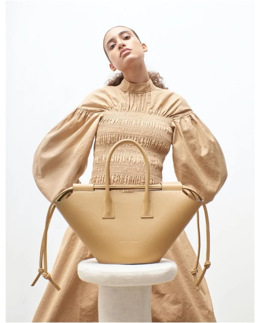 Tan-colored leather handbag sits on a white stool, woman with tan top and dress poses behind it in a studio setting, neutral lighting.