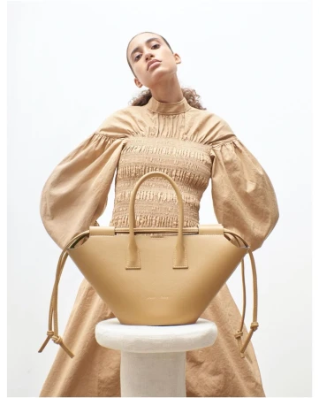 Tan-colored leather handbag sits on a white stool, woman with tan top and dress poses behind it in a studio setting, neutral lighting.