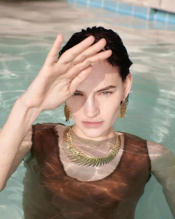 Woman in a pool wearing gold statement necklace and earrings, shielding eyes with hand, photographed with shallow depth of field.